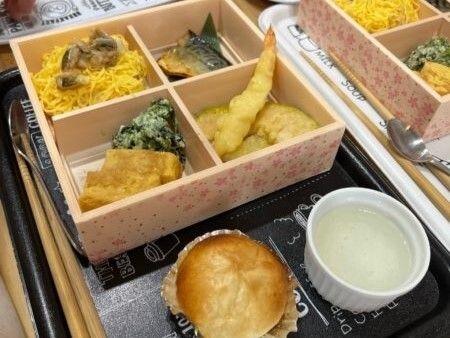 天ぷらやちらし寿司、焼き魚、煮物などが詰められた弁当風の入れ物。箸が添えられている。