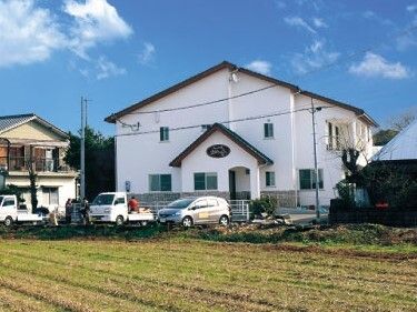 青空の下に建っている２階建ての建物は白い外壁で一般の住宅と同じような外観。畑が周りにある。