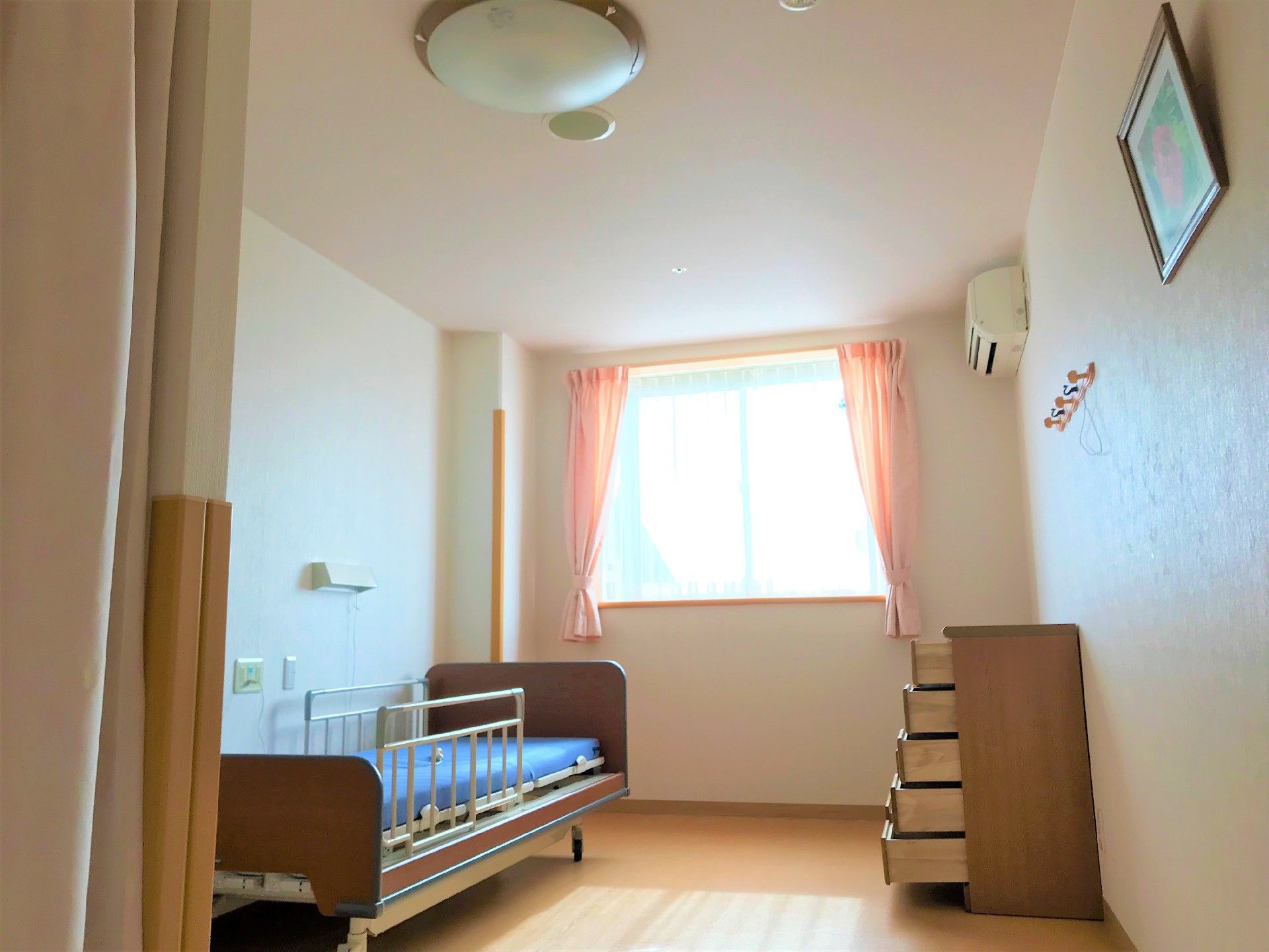 窓から日差しが差し込むエアコン付きの居室。ベッドとタンスが設置されている。