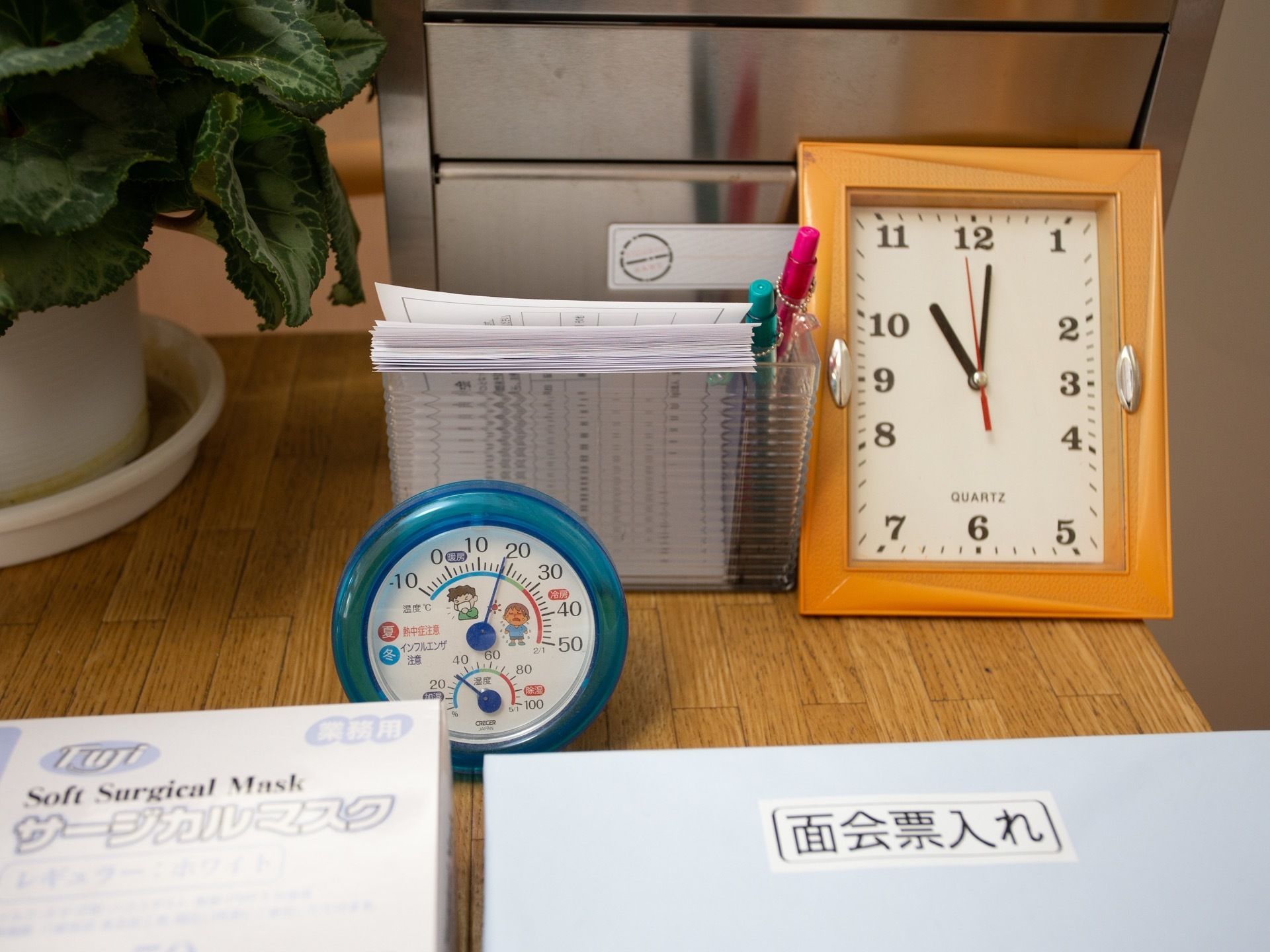玄関ホールの事務室前の台の上に、面会票、時計、温度計、マスク、消毒液などが置かれている。横に、シクラメンの鉢が飾られている。