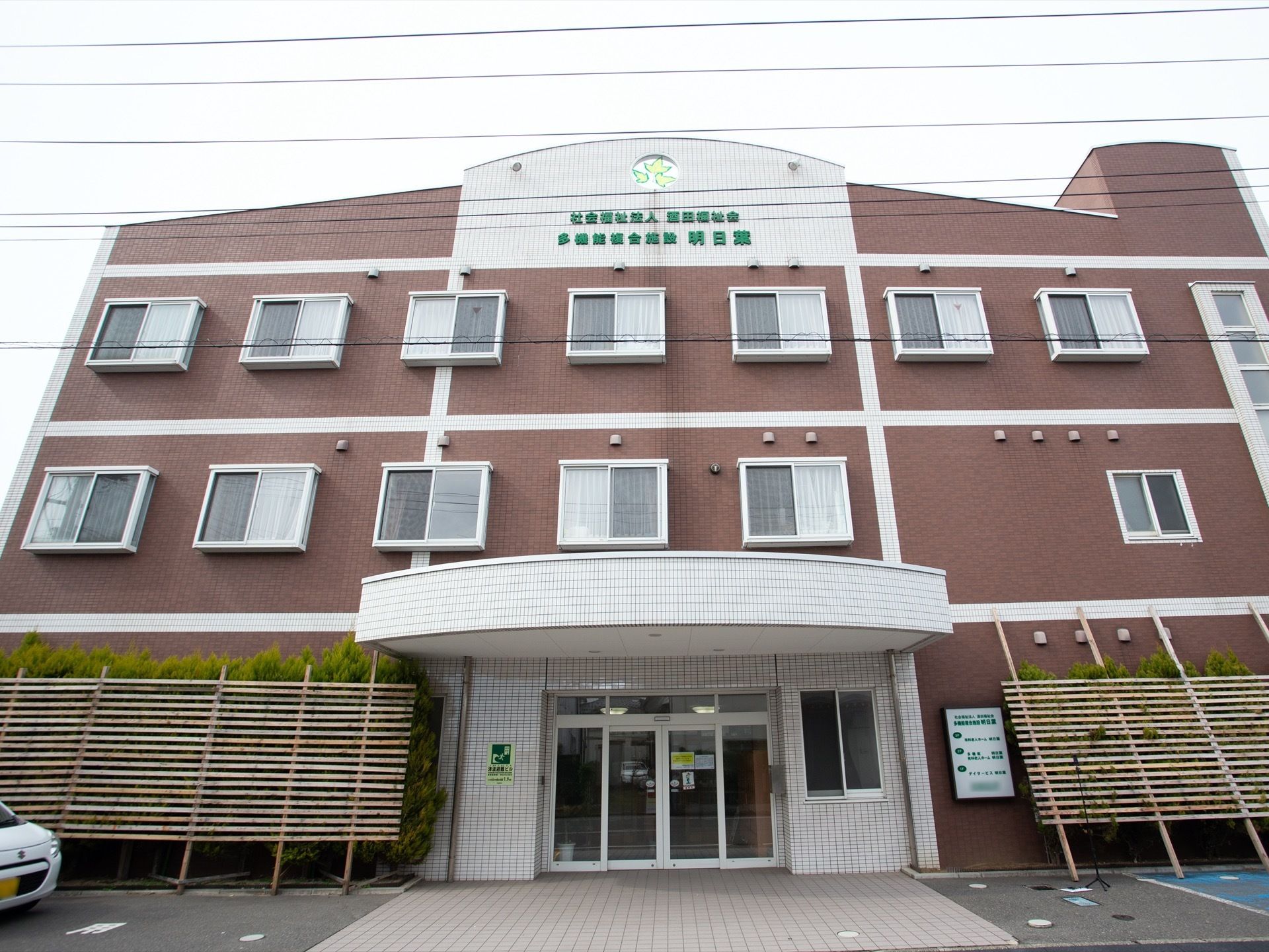 白と茶色を基調としたシンプルな外観。玄関外の屋根は大きく、扉は自動ドアとなっている。花壇には木が植えられている。