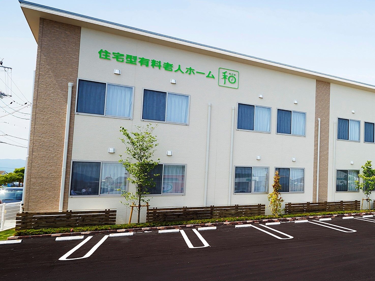 駐車場が付いた2階建ての建物。茶とベージュで家庭的な雰囲気が漂い、建物周辺には木もある。