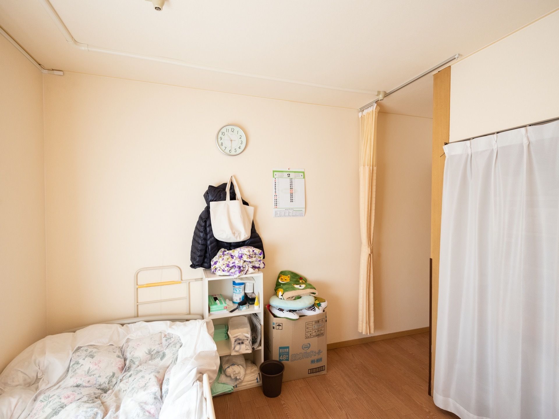 居室はホワイト色の壁紙で、壁に時計が取り付けられている。床に収納ボックスがあり、ごみ箱が置かれている。