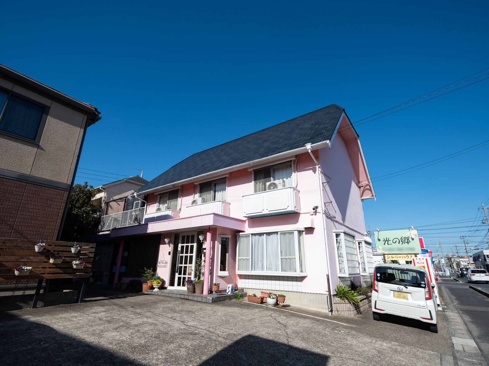 施設の建物は２階段で、ピンク色の外壁となっている。道路沿いには看板があり、ライトが取り付けられている。