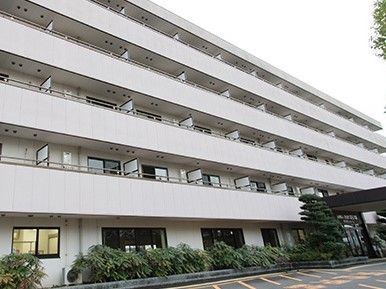 白を基調とした５階建ての建物の周りには緑が多く、玄関先には屋根が付いている。