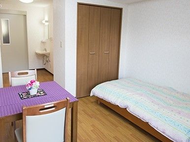 ベッドには布団がセットされており、ダイニングテーブルには紫のクロスと花が添えられ、収納スペースもある。