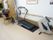 複数のリハビリ機器が並んだリハビリスペースでいすに座りコードを引っ張っている高齢女性。