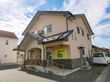 ベージュの2階建ての建物にはグレーの瓦屋根が付き、建物の玄関には手すり付きの階段や屋根があり、駐車場もある。