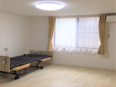 エアコンが付いたフローリングの部屋にはカーテンが付いた窓や介護用ベッドがあり、ベッドサイドには緊急通報装置もある。