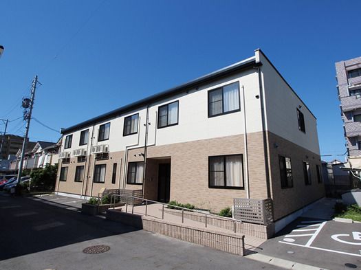 2階建ての建物は白とベージュでフロアにより外壁の色が異なる。駐車スペースがあり、玄関には手すり付きのスロープがある。