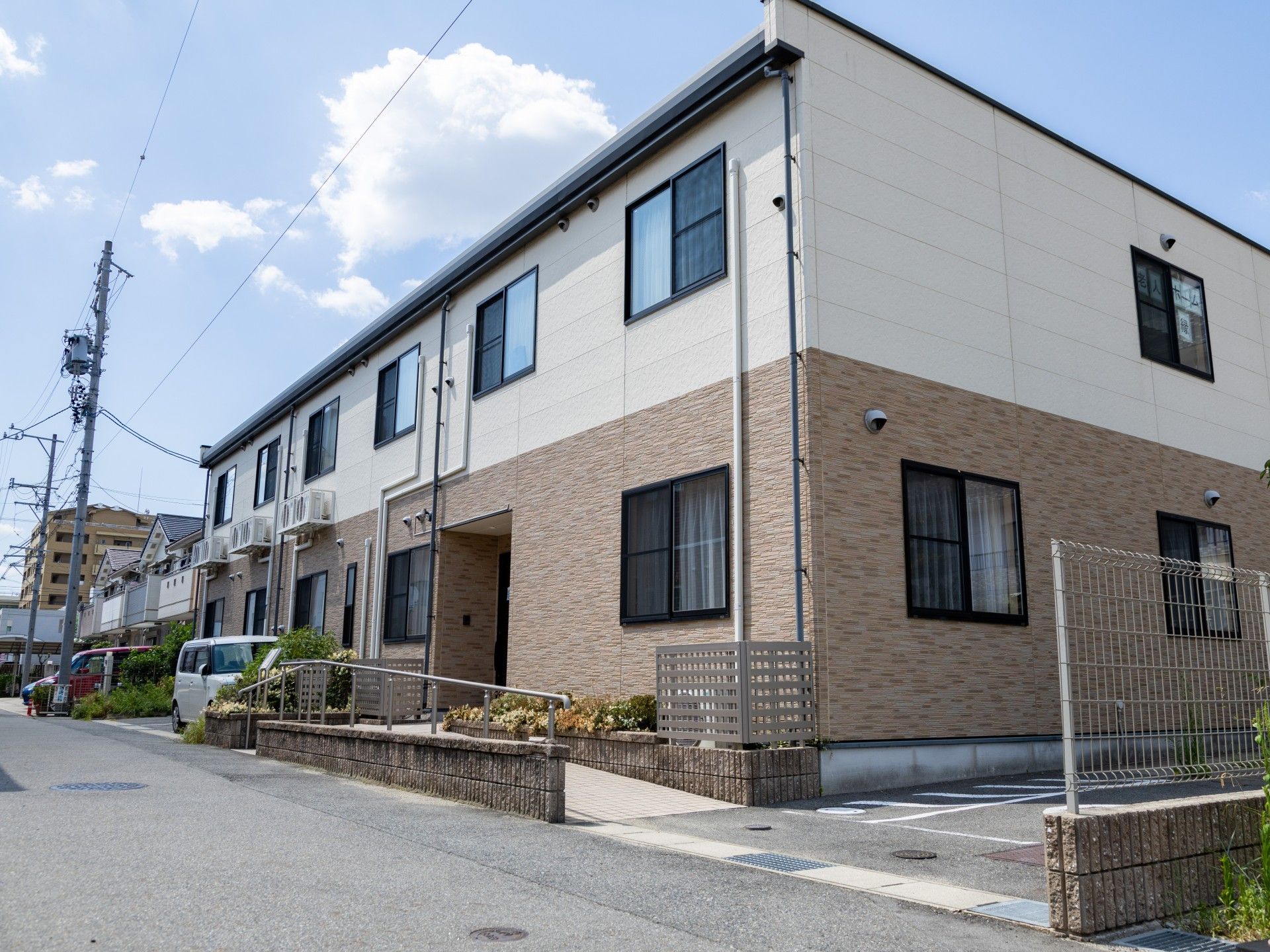駐車場が付いた2階建ての建物はフロアにより外壁の色が異なり、住宅地の中にある。玄関には手すり付きのスロープが付いている。