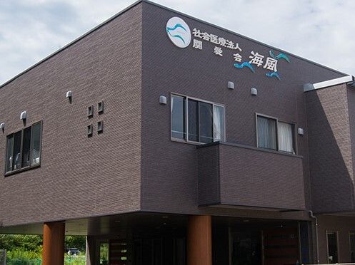 ブラウン系の建物の外壁にはホーム名が書かれ、周辺には緑が多い。日当たりのいい場所に建っている。