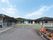平屋建ての建物が、コの字型に立っており、白い柵を隔てて手前に駐車場がある。柵の中央に、水色の看板が立っている。