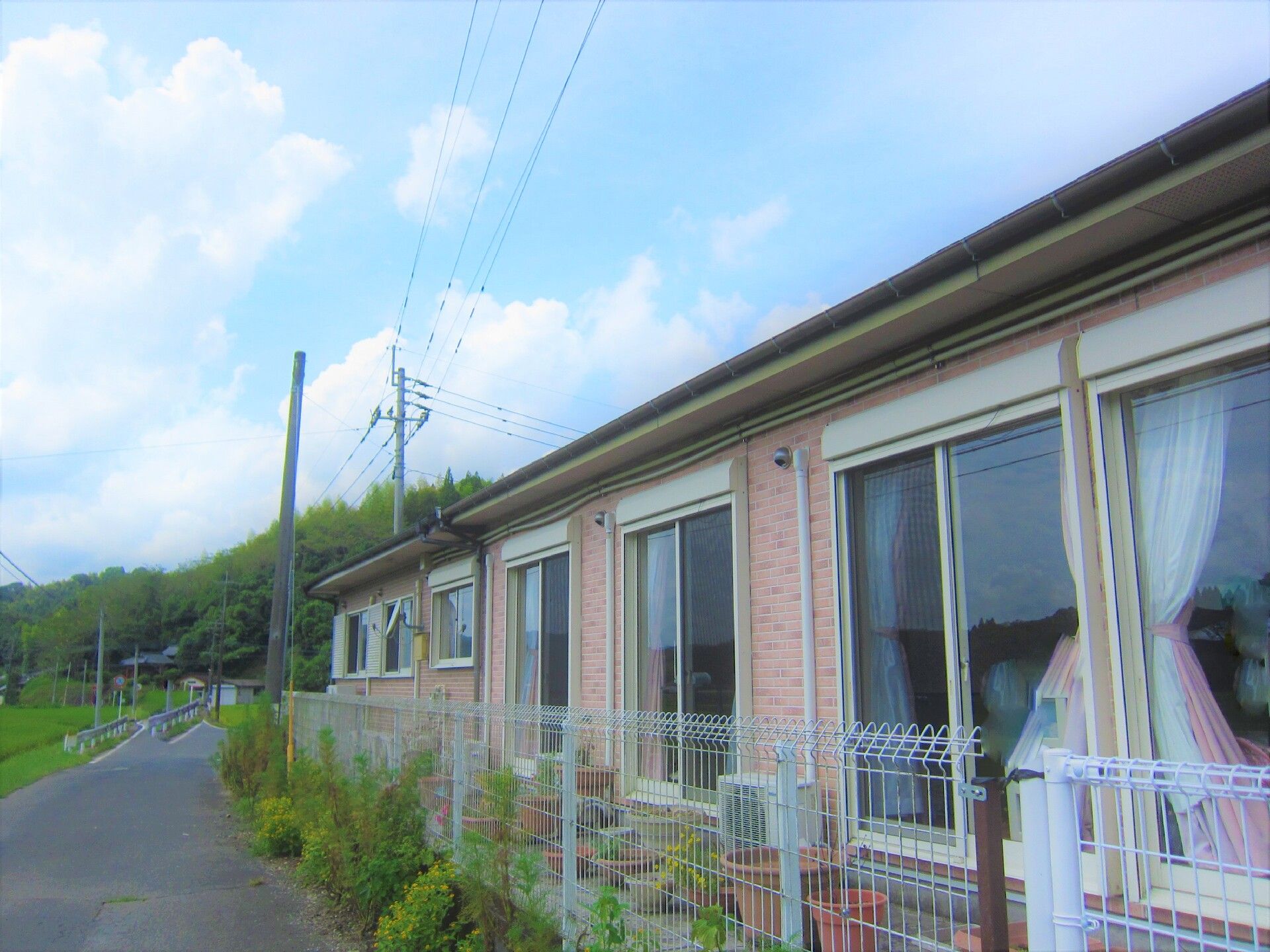 カーテンが付いた大きな窓がたくさんついた平屋建ての建物。周りには白いフェンスが建てられ、周辺には自然が多い。