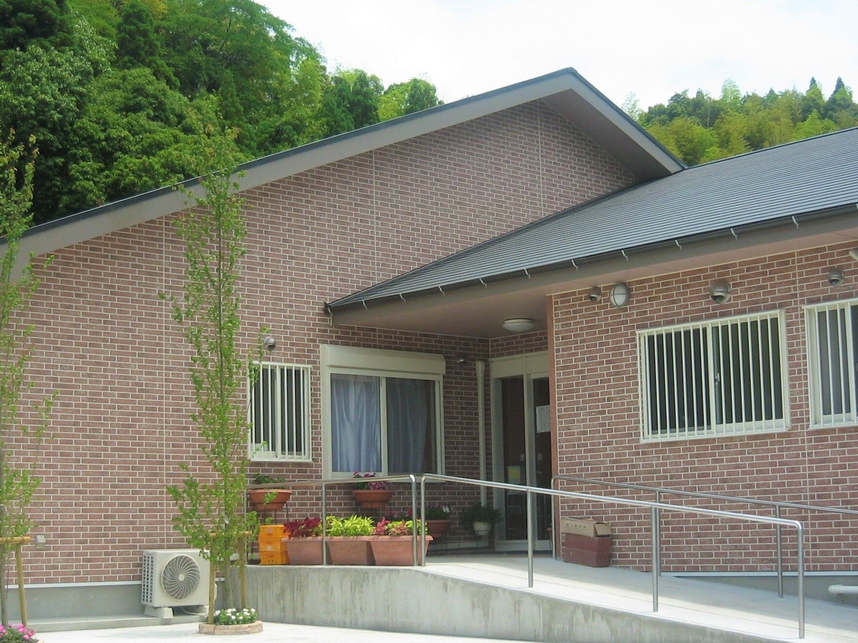 三角屋根が付いたブラウン系の建物は平屋建て。周辺には緑が多く、敷地内には花が植えられたプランターが置かれている。