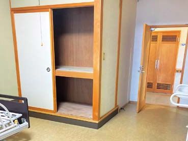 オレンジのカーディガンを着た眼鏡姿の高齢女性が車いすに座り、白衣を着た女性スタッフが笑顔で押している。