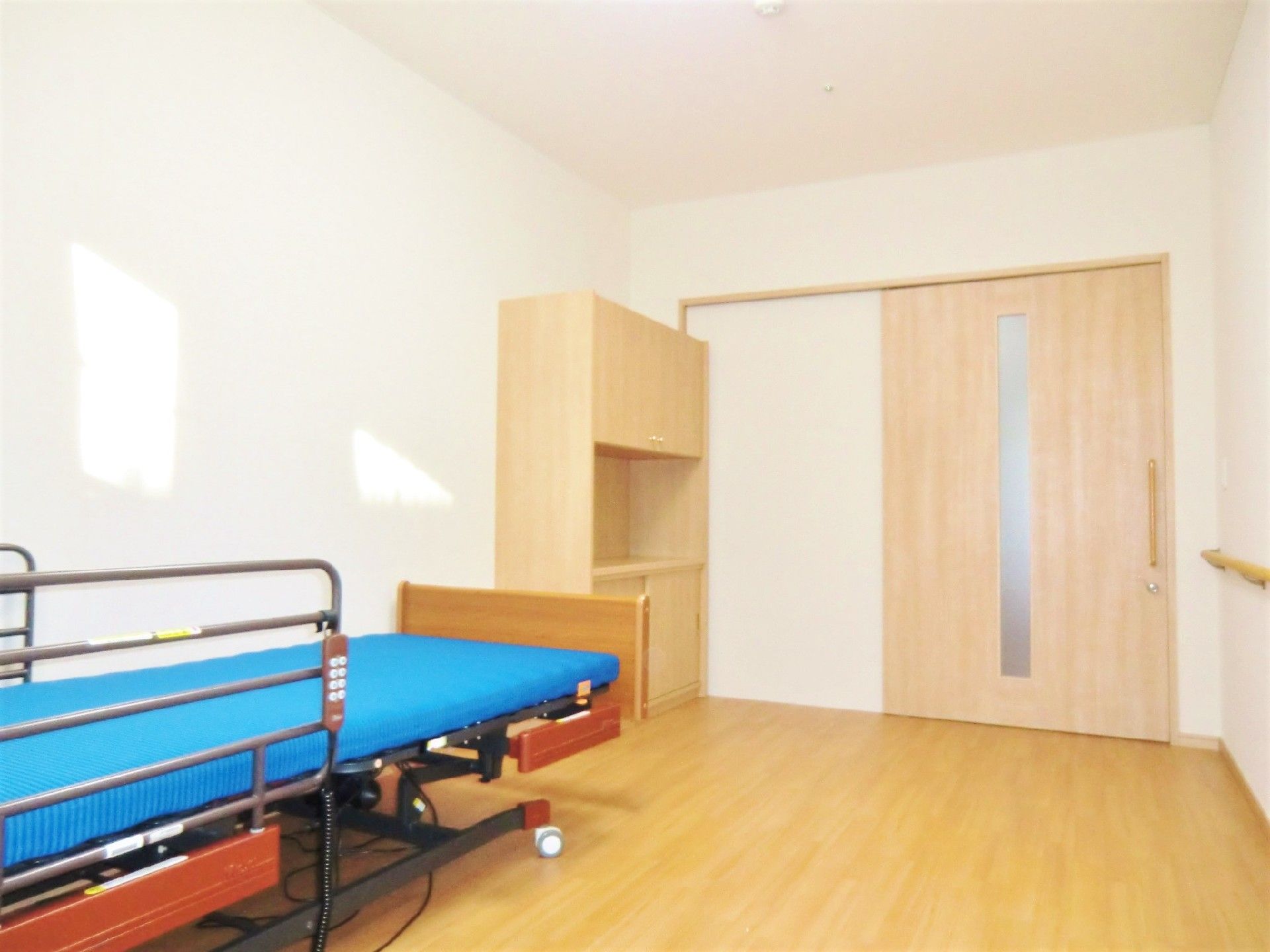 小さなベッドと物入が置いてある施設内の居室。右側奥に引き戸仕様のドアがあるフローリングの部屋