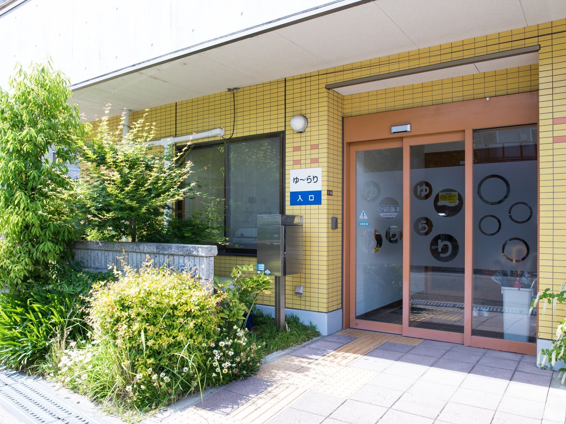 黄色とオレンジのタイルが貼られた玄関である。入口前にはたくさんの植栽が植えられている。扉の前にポストがある。