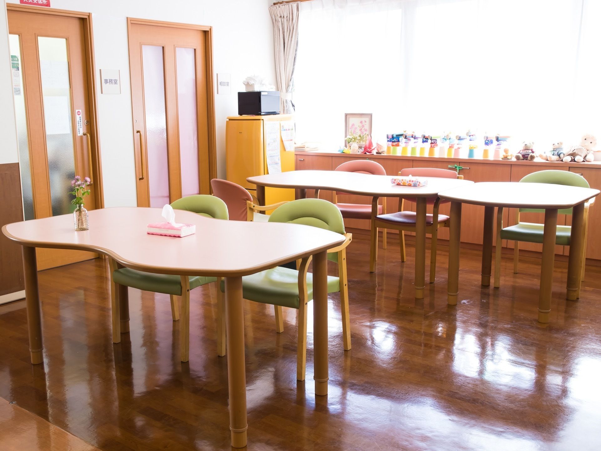 テーブルセットが並べられた明るい部屋がある。窓際の棚にはぬいぐるみや入居者様の作った作品が飾られている。