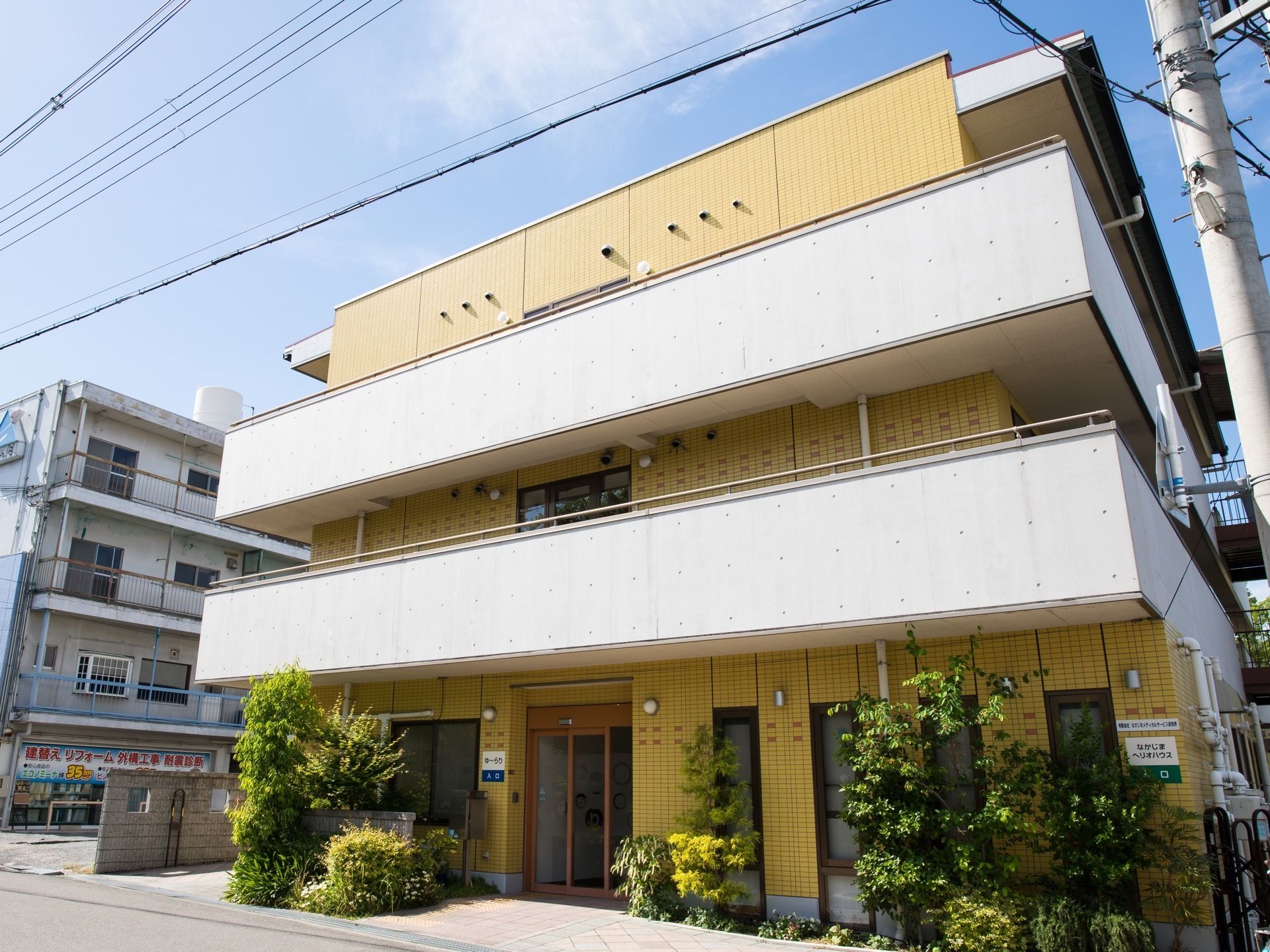 黄色いタイルと打ちっぱなしのコンクリートが印象的な3階建ての建物がある。玄関付近には緑が植えられている。