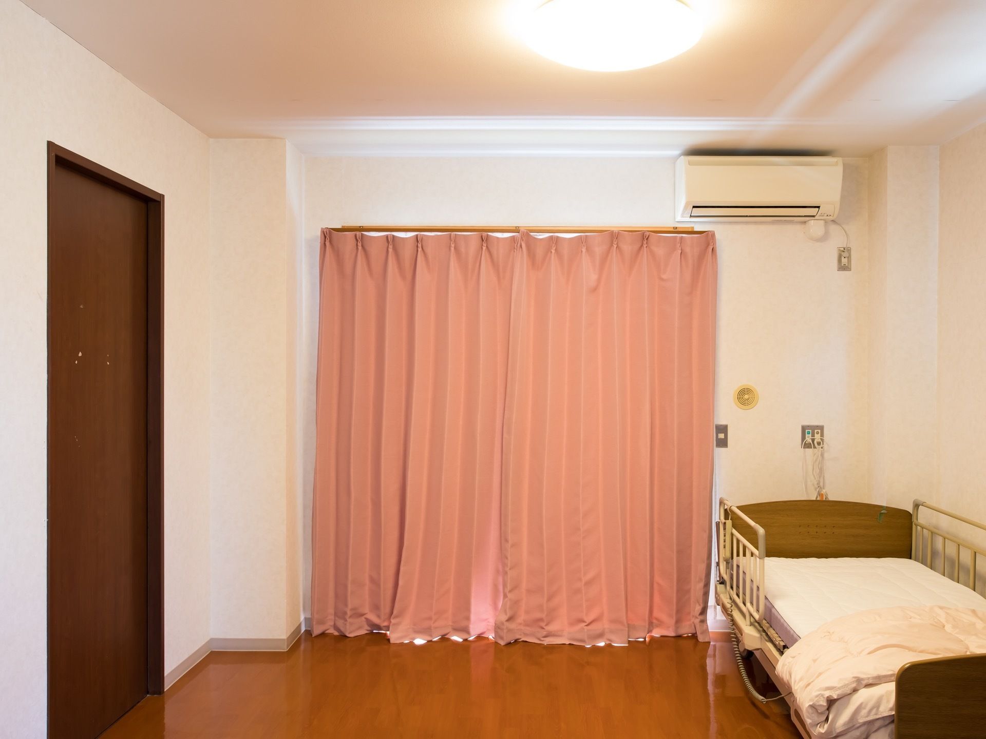 フローリングの部屋にベッドが一台置かれている。掃出しの窓にピンク色のカーテンが掛けられ、その横にはエアコンがついている。