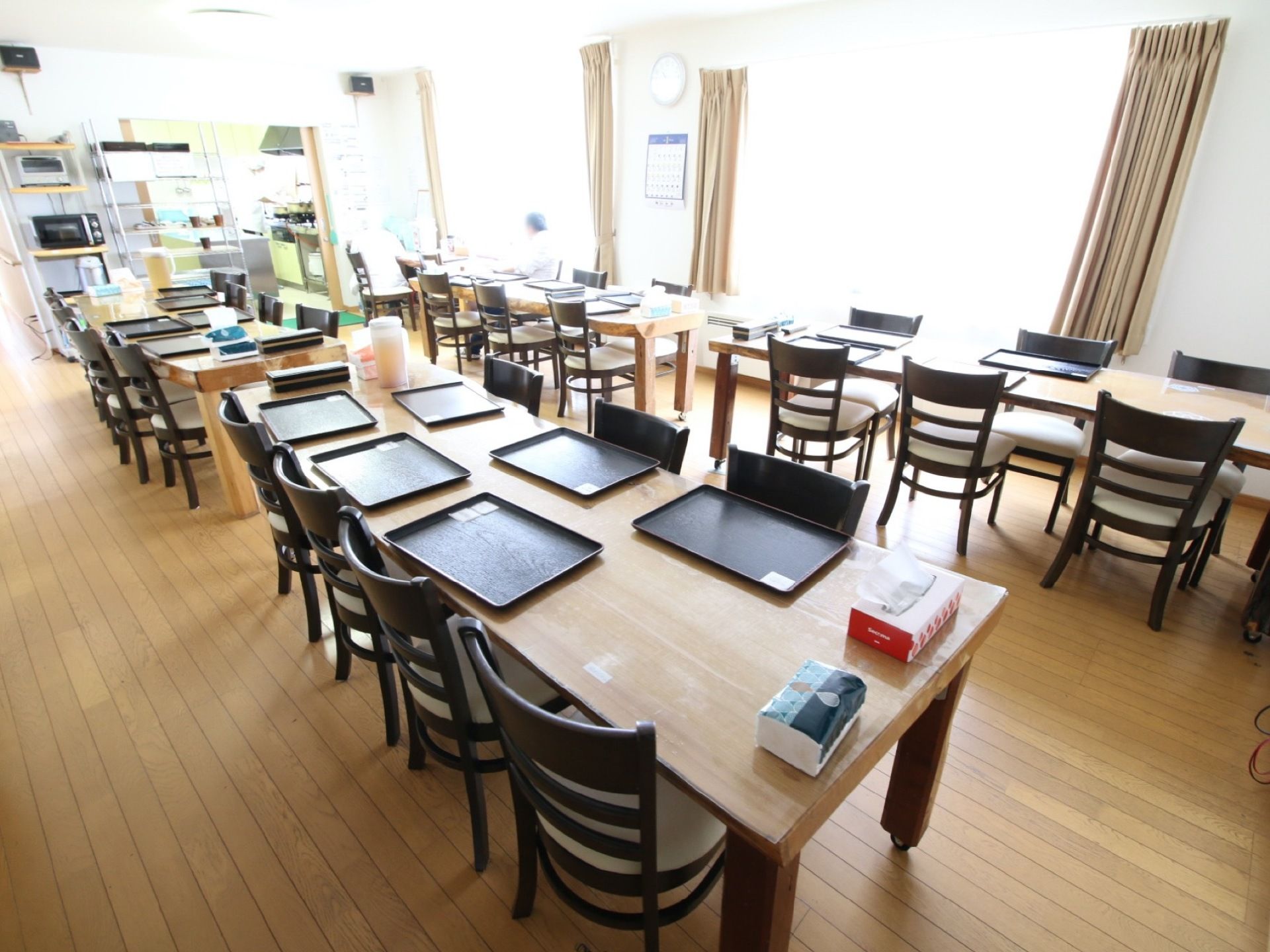 大きな窓から日射しが入る環境のよい食堂である。ダイニングテーブルが横に２列並んでいる。電子レンジが２台完備されている。