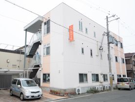 白を基調とした3階建ての建物になっている。道路に車が1台停まっており、駐車スペースにも1台車が停められている。