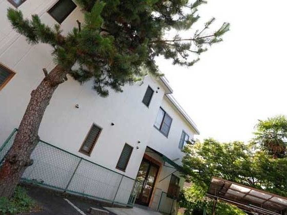 白い外壁の２階建ての建物は日当たりが得、周辺にはフェンスがあり、木もたくさん生えている。