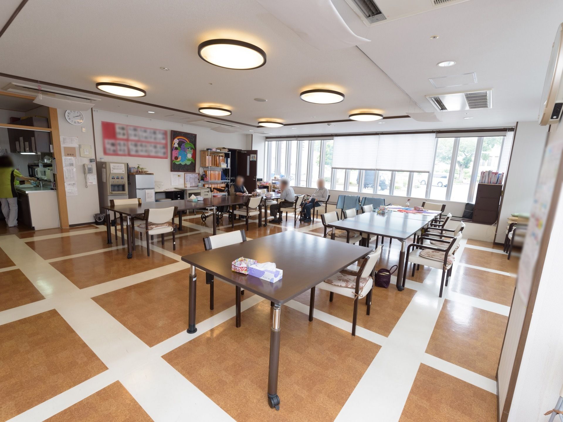 窓側のテーブル席に3名の入居者様が座って談笑されている。壁に書道やちぎり絵などの作品が綺麗に展示されている。