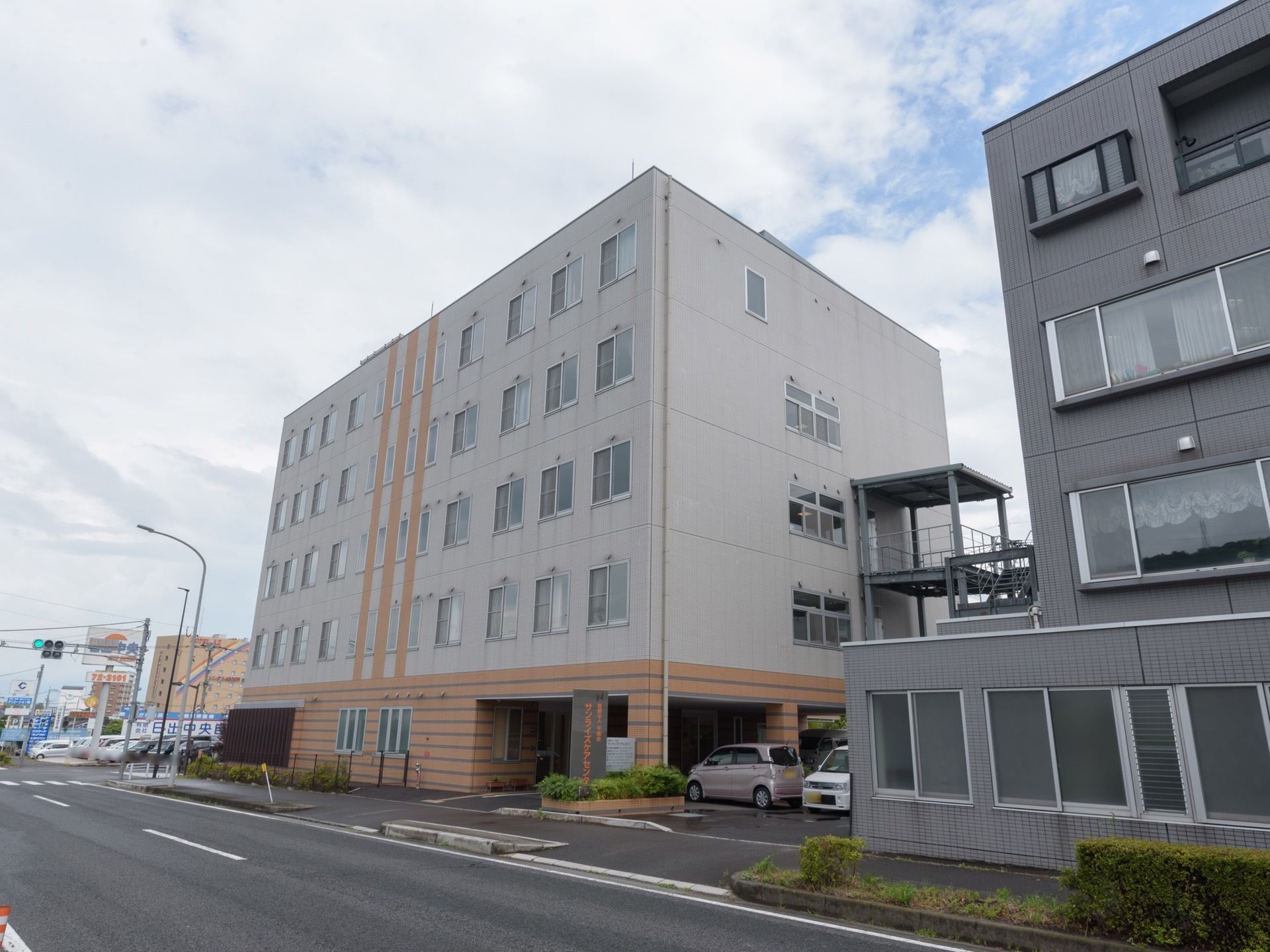 5階建ての白とオレンジの外壁の建物で、大きな道路に面して建てられている。ひさしの下に数台の車が停車している。