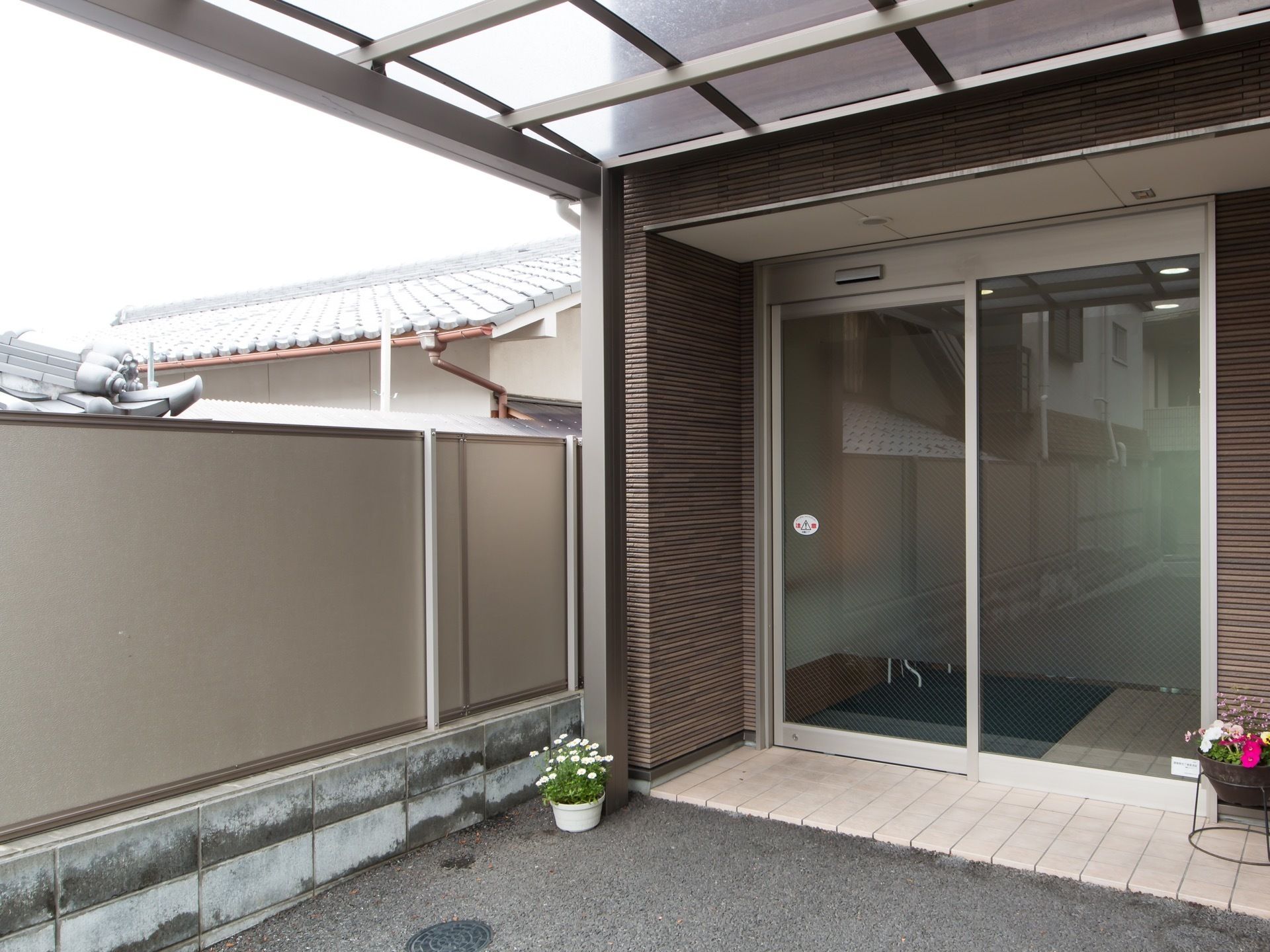 自動ドアの玄関で上部には大きなひさしが設けられている。地面には花が植えられた植木鉢が2つ置かれている。