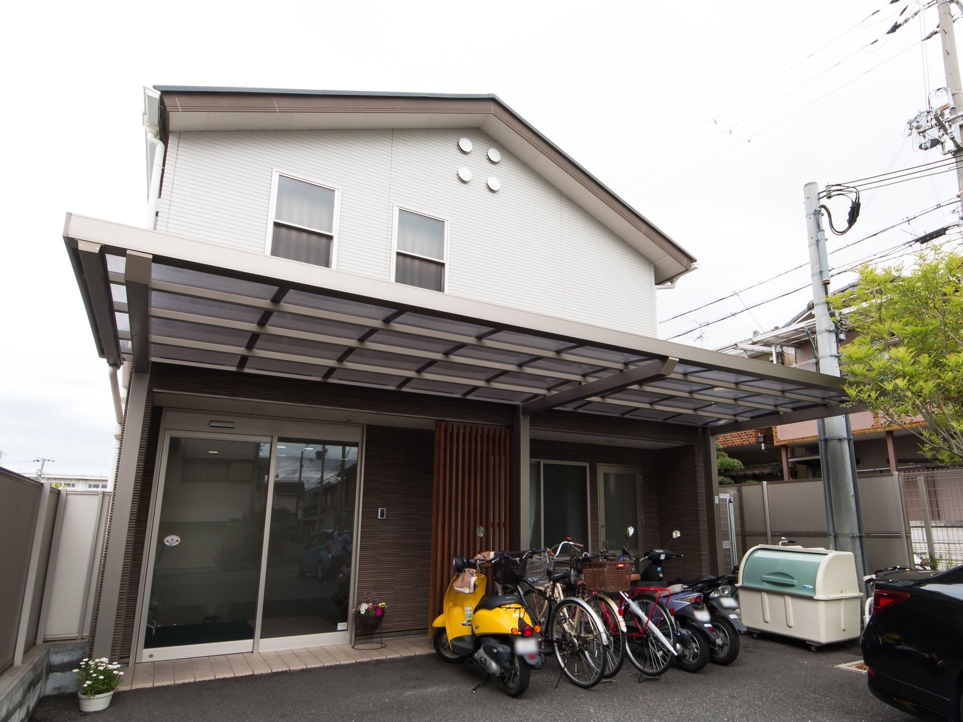 大きなひさしが設置された玄関で、自動ドアの右側にはバイクや自転車が停められている。自転車の手前側に車が1台ある。