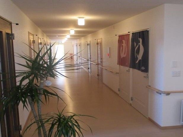 白髪の高齢女性が座っていて、紺のポロシャツを着た女性スタッフが後ろから肩に白いタオルをかけ微笑みかけている様子。