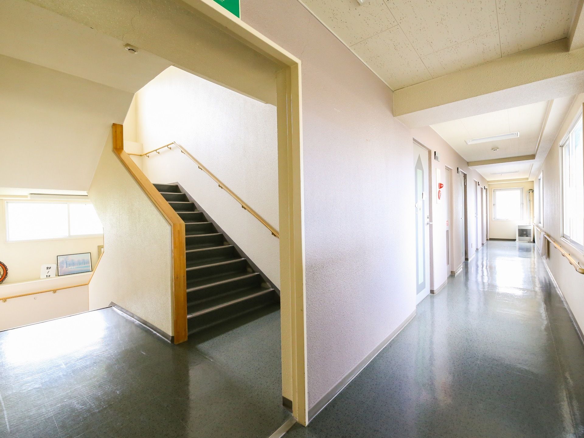 まっすぐに伸びている廊下の右側の壁には窓が連続していて、その下方に手すりが施されている。左側には、横幅の広い階段があるのが見える。