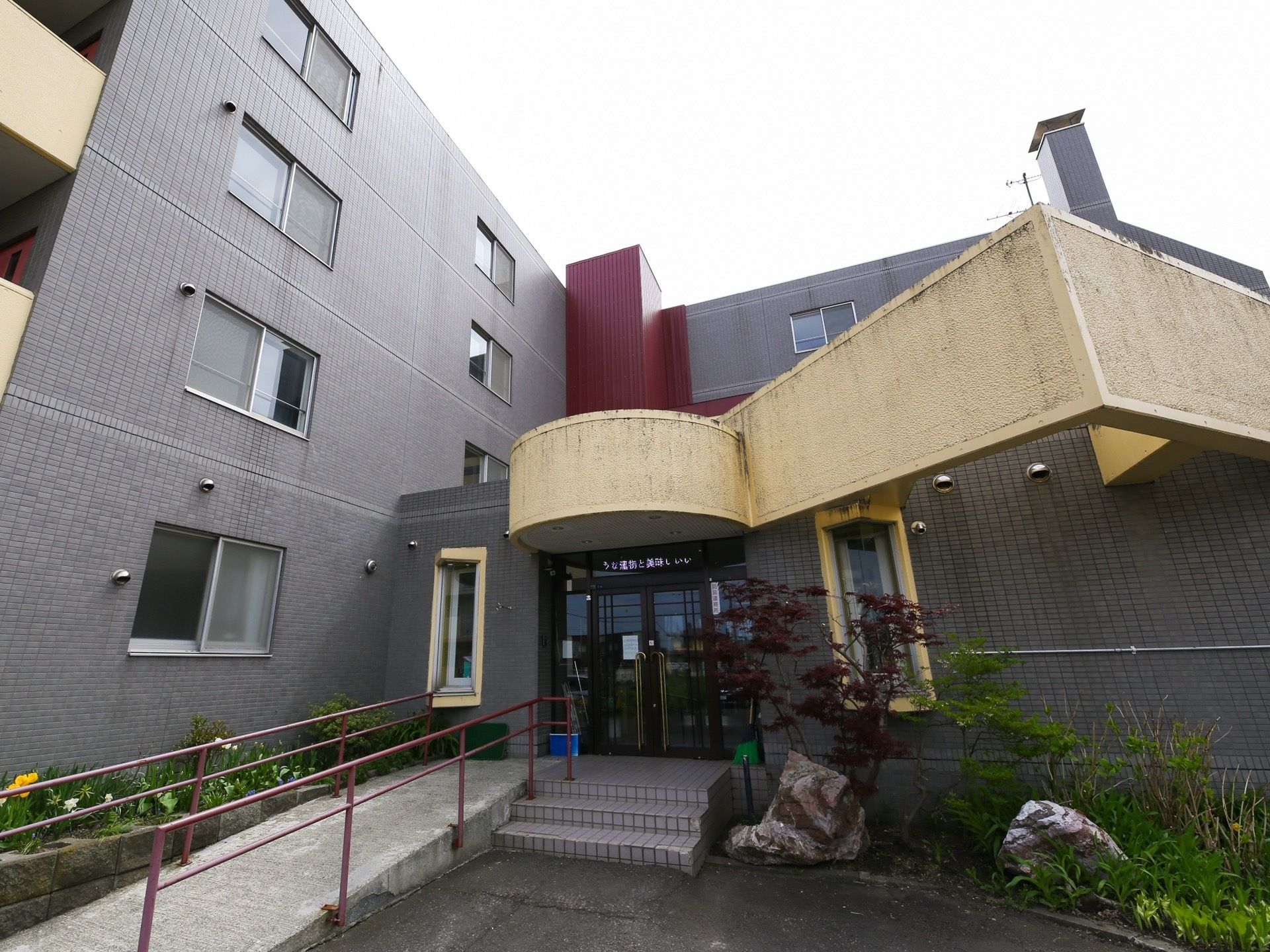 玄関の前面には、階段とスロープが設置されている。建物の周囲には、植栽が施されており、大きな石も配されている。