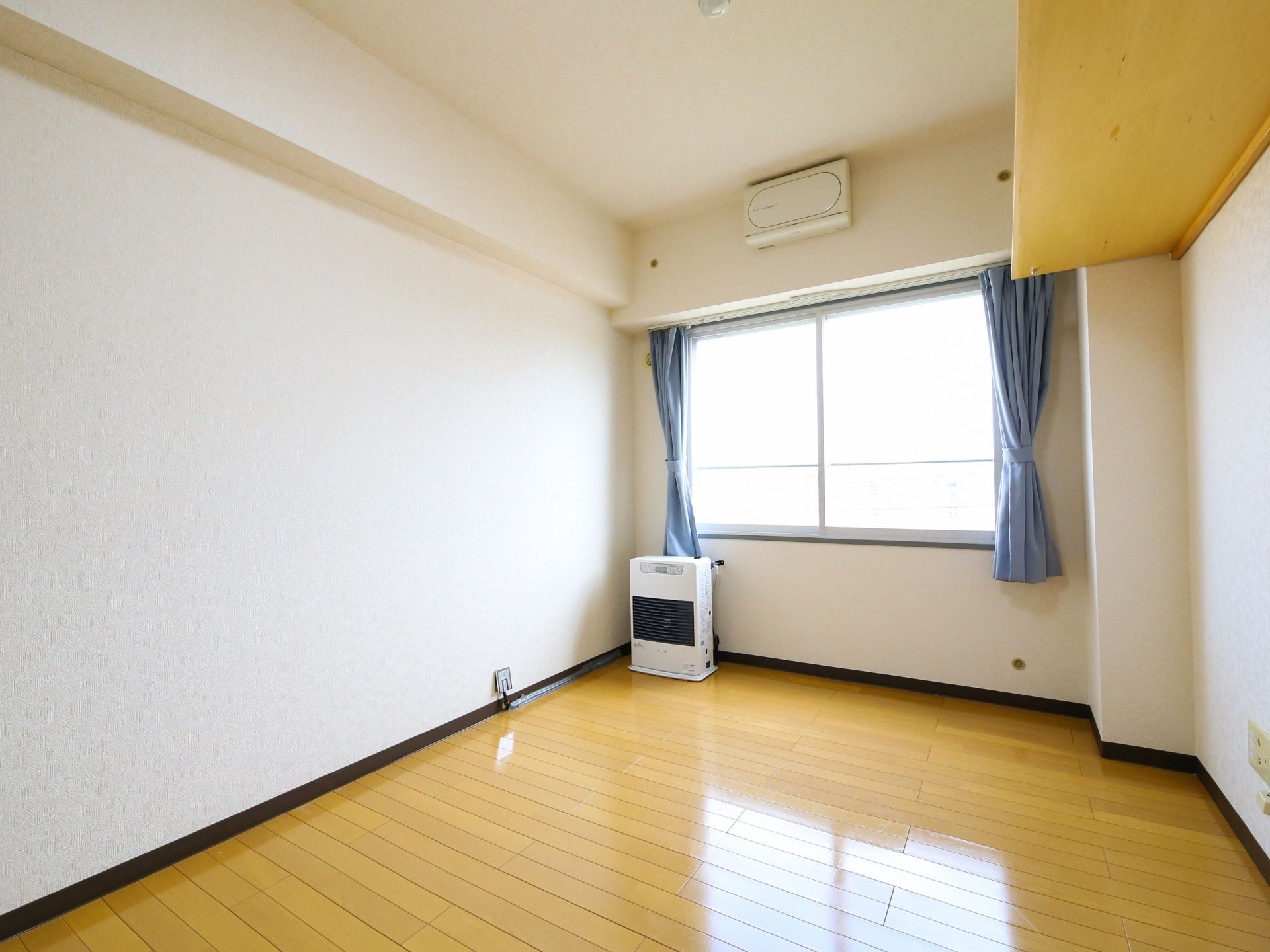 窓側にストーブが置かれているワンルームタイプの居室である。右側の壁面の上方に棚板が取りつけられている。