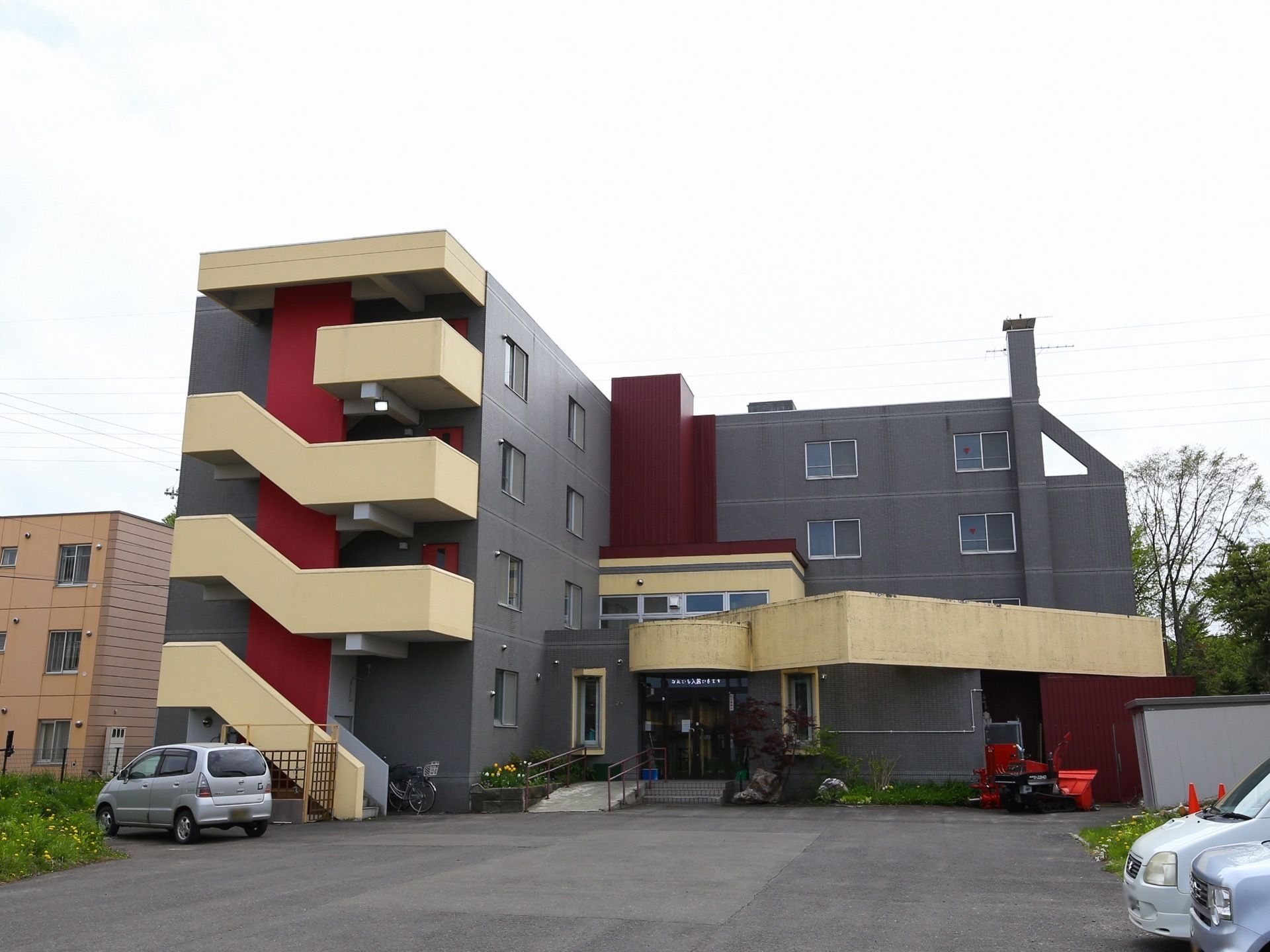 グレーにブラウンの切り替えのある外壁で、左手前にベージュの外階段がある4階建ての建物