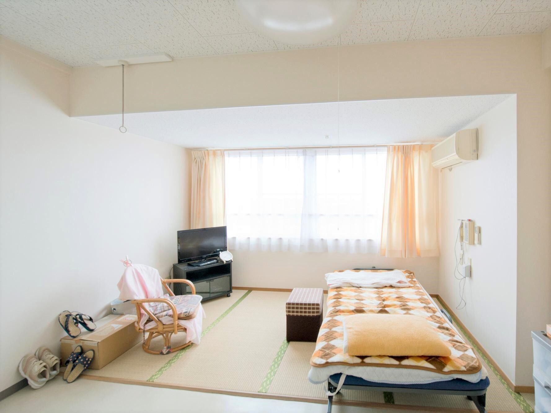 エアコンが付いたシンプルな居室。大きな窓のそばにはテレビもあり、ベッドやいすもついている。