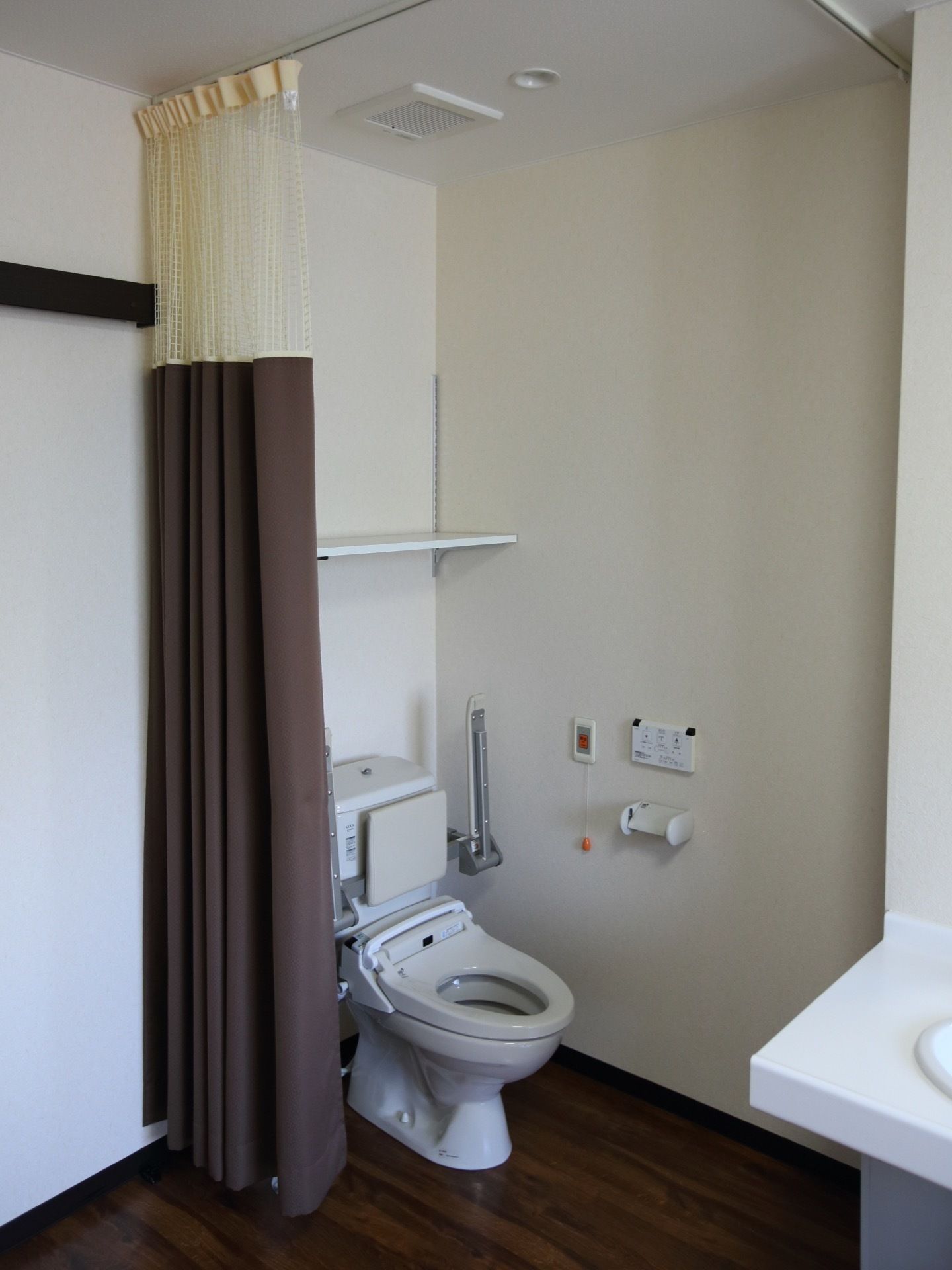 居室に備え付けられているトイレである。便器には背もたれやひじ掛けが設置されており、上部には収納棚が設けられている。
