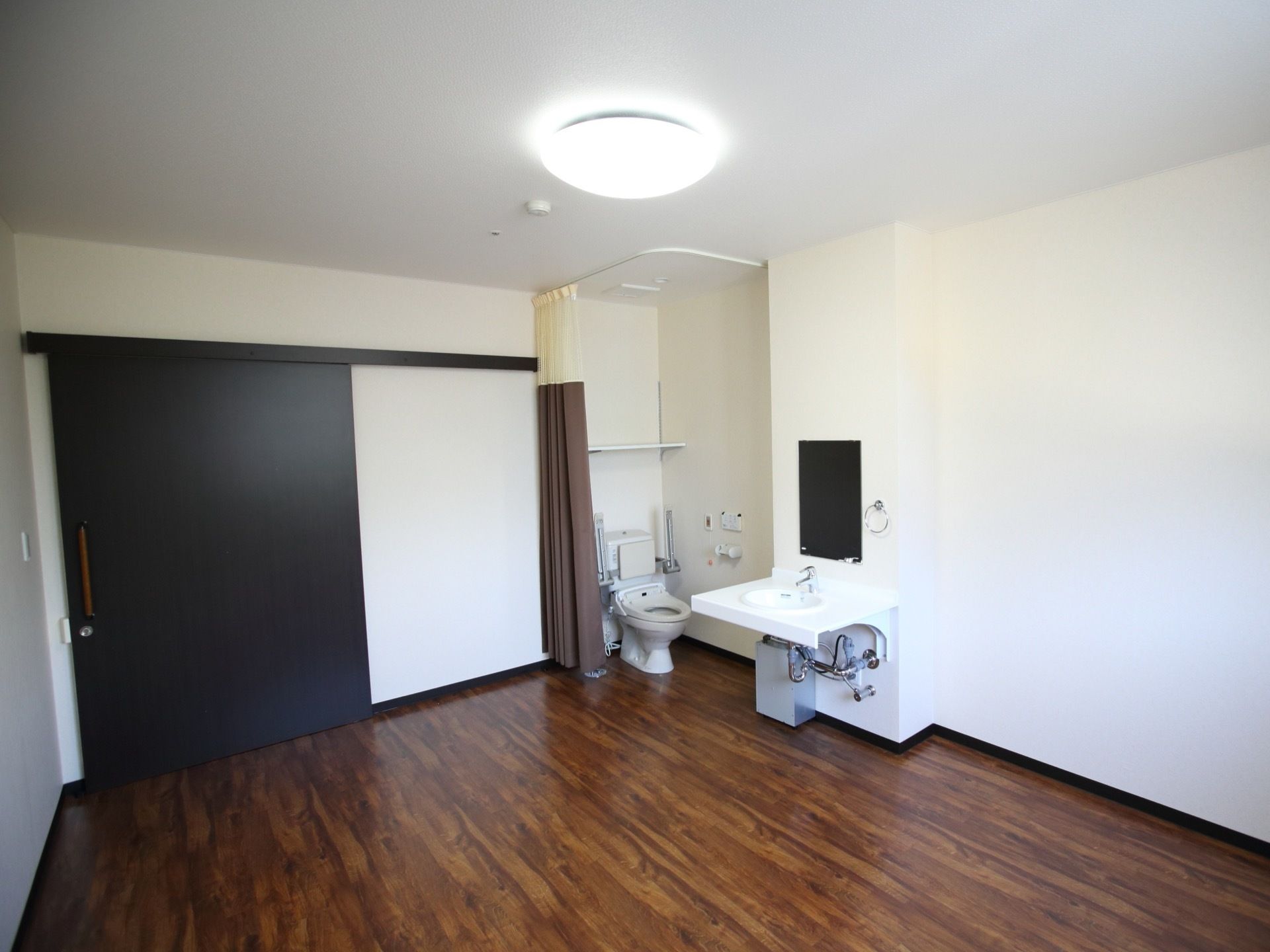 洗面台やトイレが完備されている広々とした居室で、天井に照明が設置されている。入口は引き戸が採用されている。