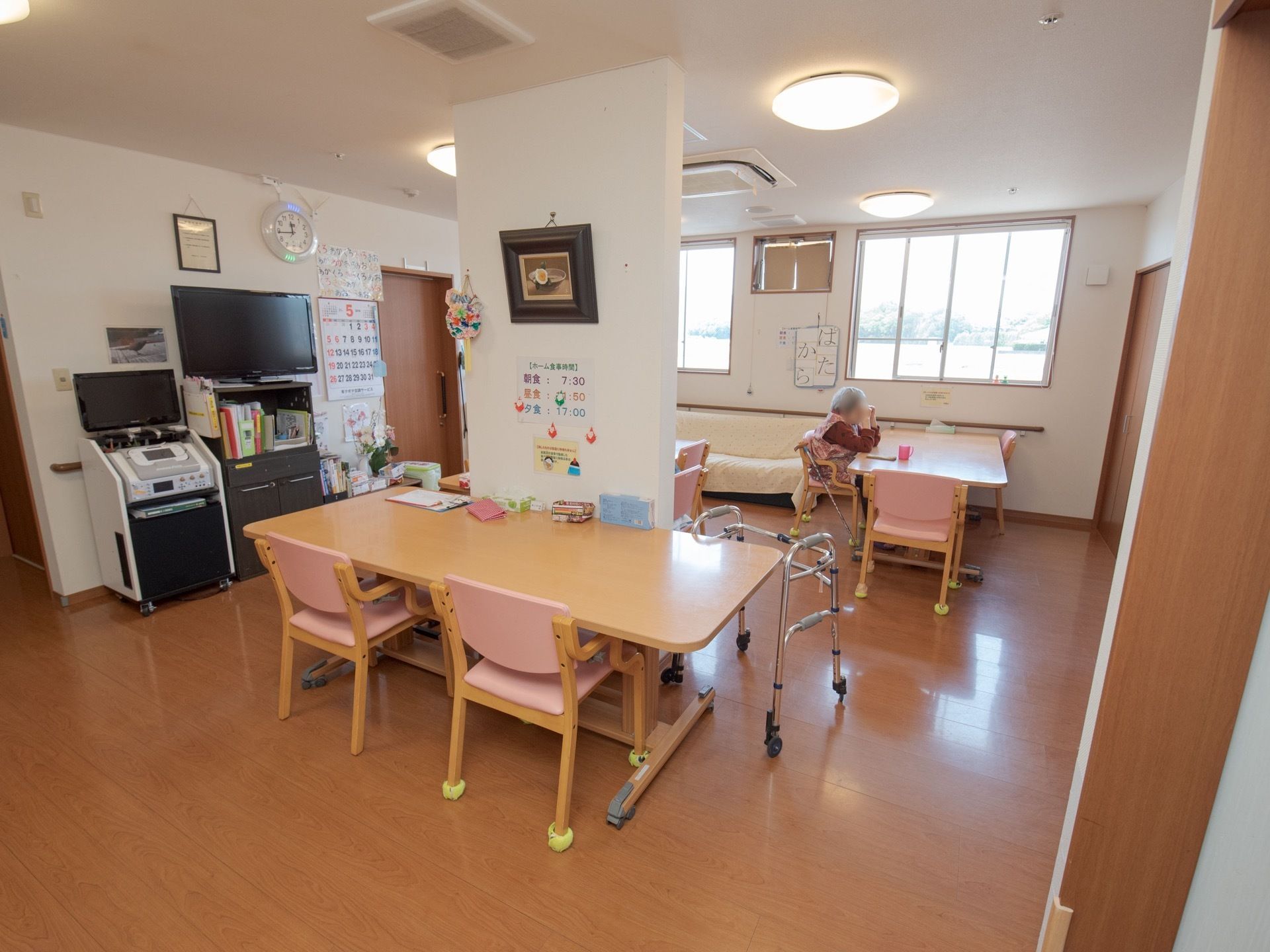 高窓から外の景色が見える。テーブルと椅子のセットが数組と窓下にはソファーが置かれている。左壁に大きなテレビと時計などが掛けられている。