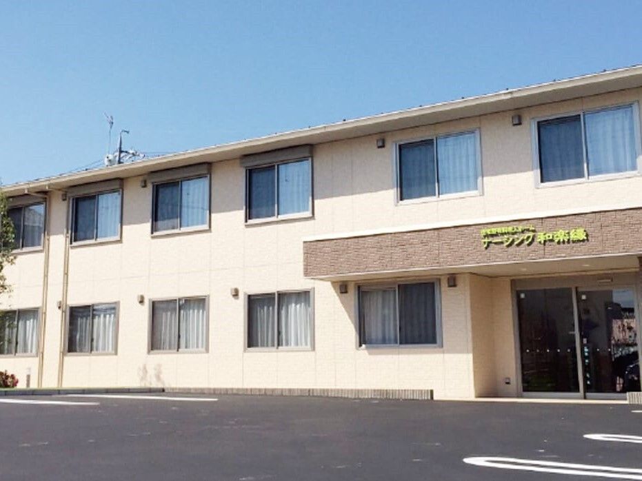 ベージュの外観の２階建ての建物は玄関に屋根があり、広い駐車場が付いている。