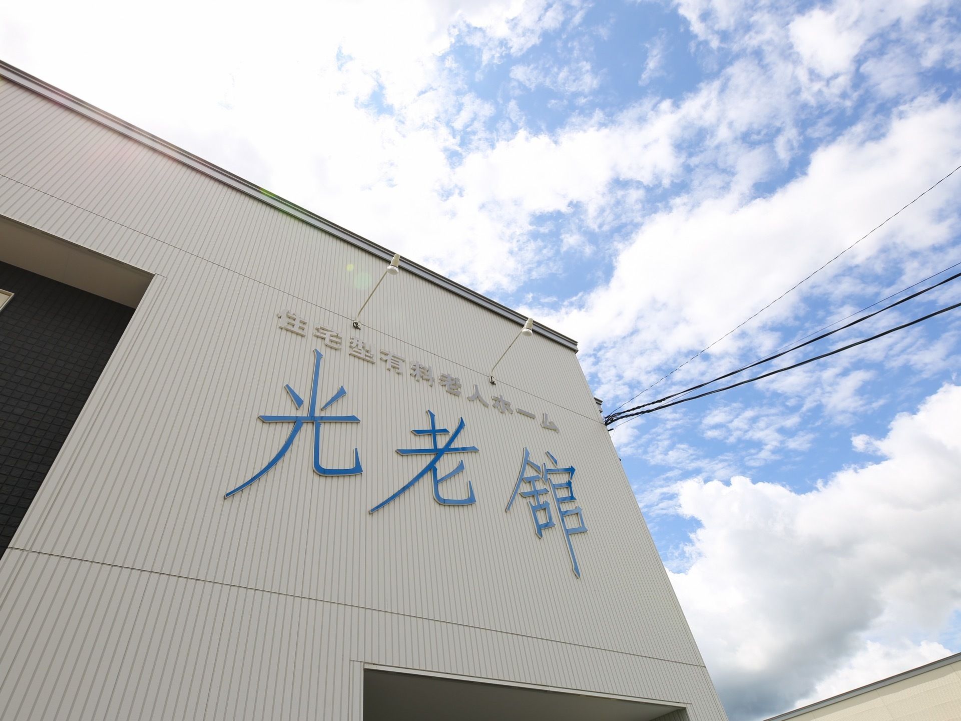 白を基調とした外壁に、青い文字で大きく施設名が書かれている。上には小さな２つの照明が設置されている。