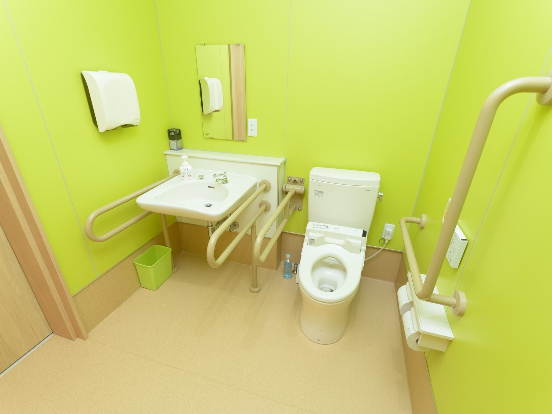 壁が若草色、床がベージュで、明るい印象のトイレである。洗面台とトイレが同一スペースに置かれ、周囲が広い。便座と洗面台には手すりが付いている。
