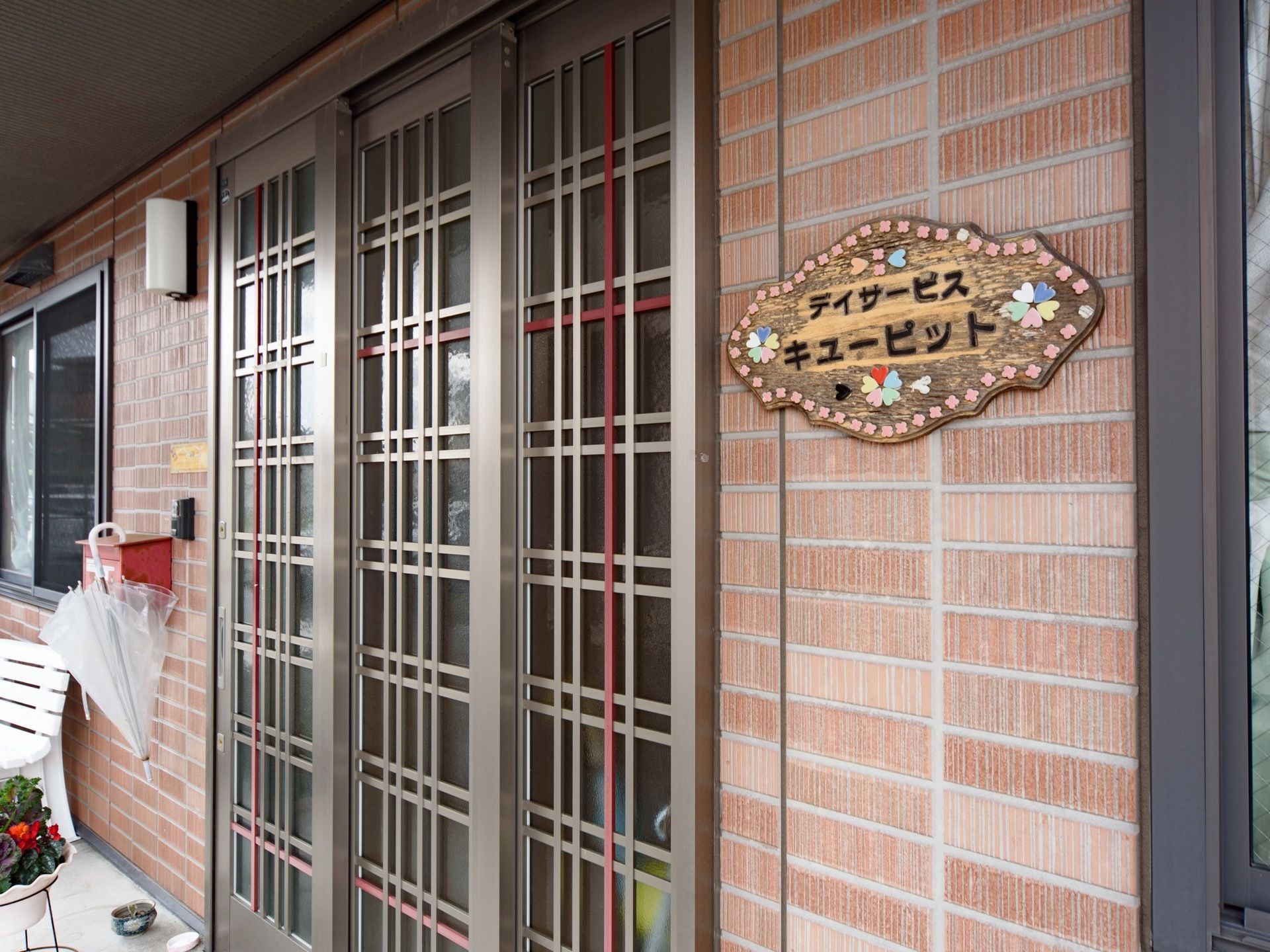 エントランスのドアには、デイサービスの銘板が設置されている。花に囲まれた木製の板に、丸みを帯びた黒い文字で施設名が表示されている。