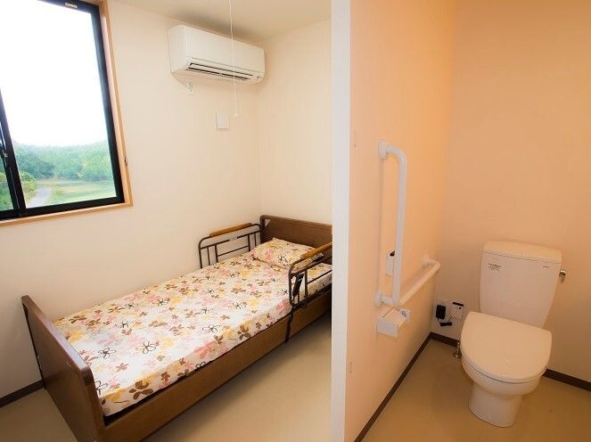 エアコンや介護ベッド、エル字型の手すりやオレンジの呼び出しボタンが付いたトイレがある個室の部屋。