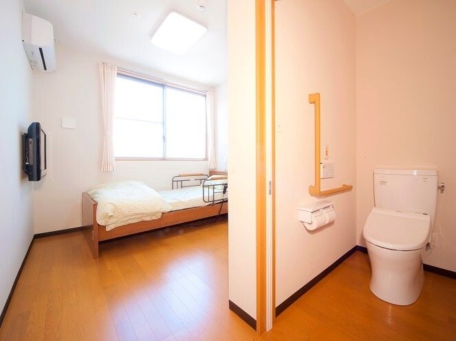 エル字型の手すりやオレンジの呼び出しボタンが付いたトイレがフローリングの部屋にあり、テレビやエアコンもある。