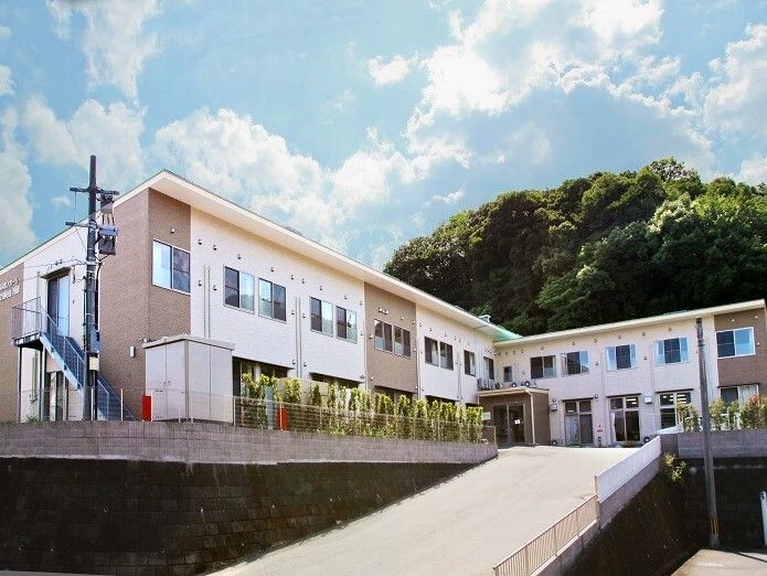 エル字型の２階建ての建物の後ろには山があり、敷地内には草木が植えられ、建物の前には坂がある。