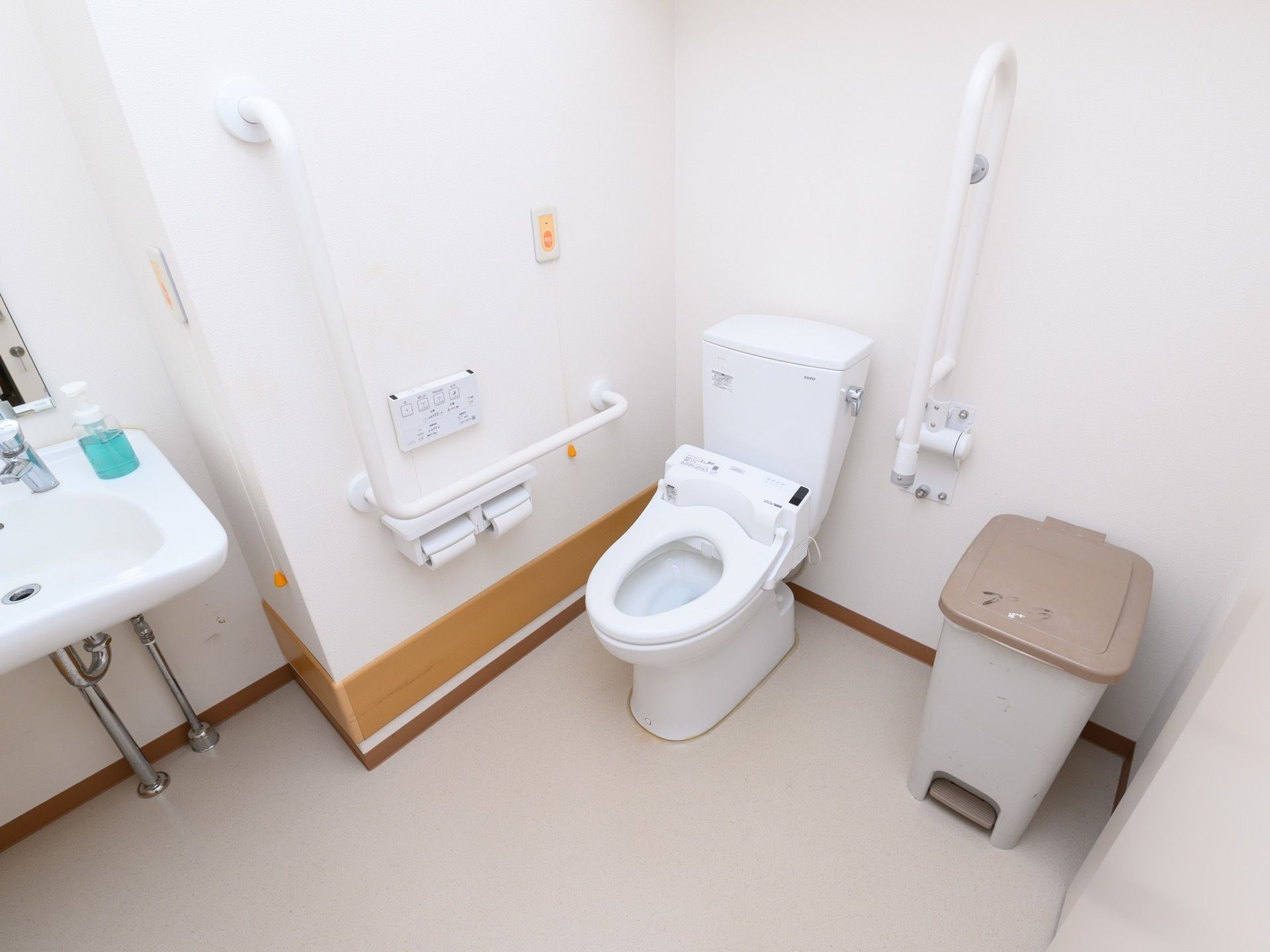 白い壁にナースコールや手すりが設置された個室トイレで、洋式便座の右側に大きめのゴミ箱が１つ用意されている。