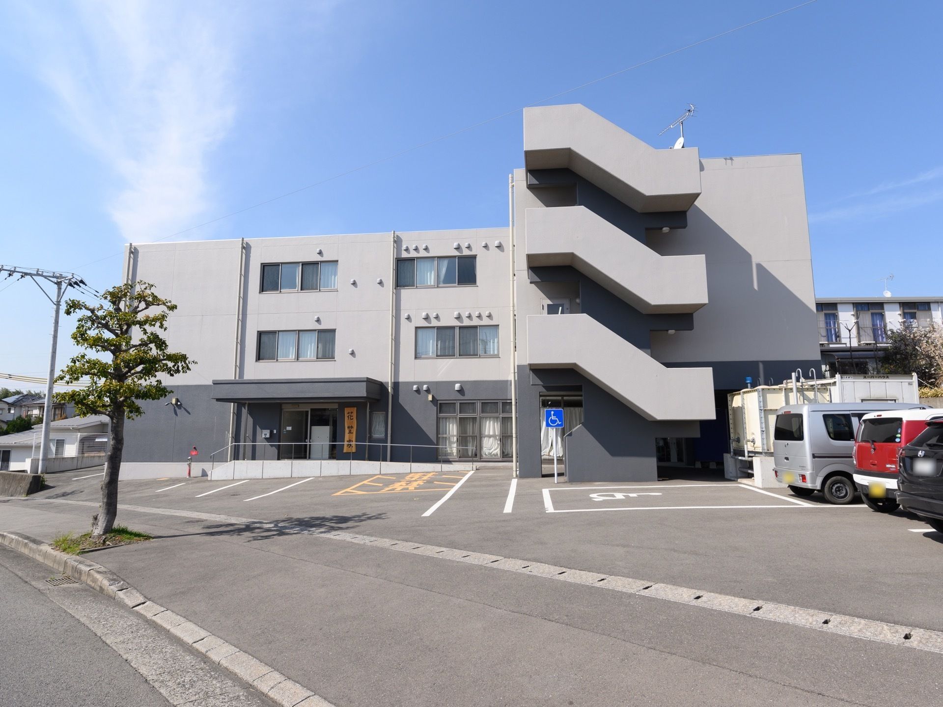 グレーを基調とした外壁の３階建ての建物で、外階段が設置されている。施設前の路面は舗装されており、車が数台停められている。